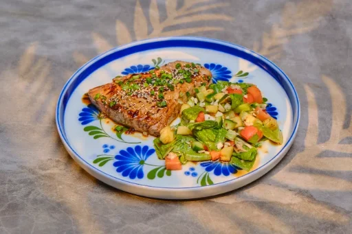 A thick piece of seared fish topped with sesame seeds and chopped green onion, served beside a salad of lettuce, cucumber and tomato on a white plate with blue flowers.